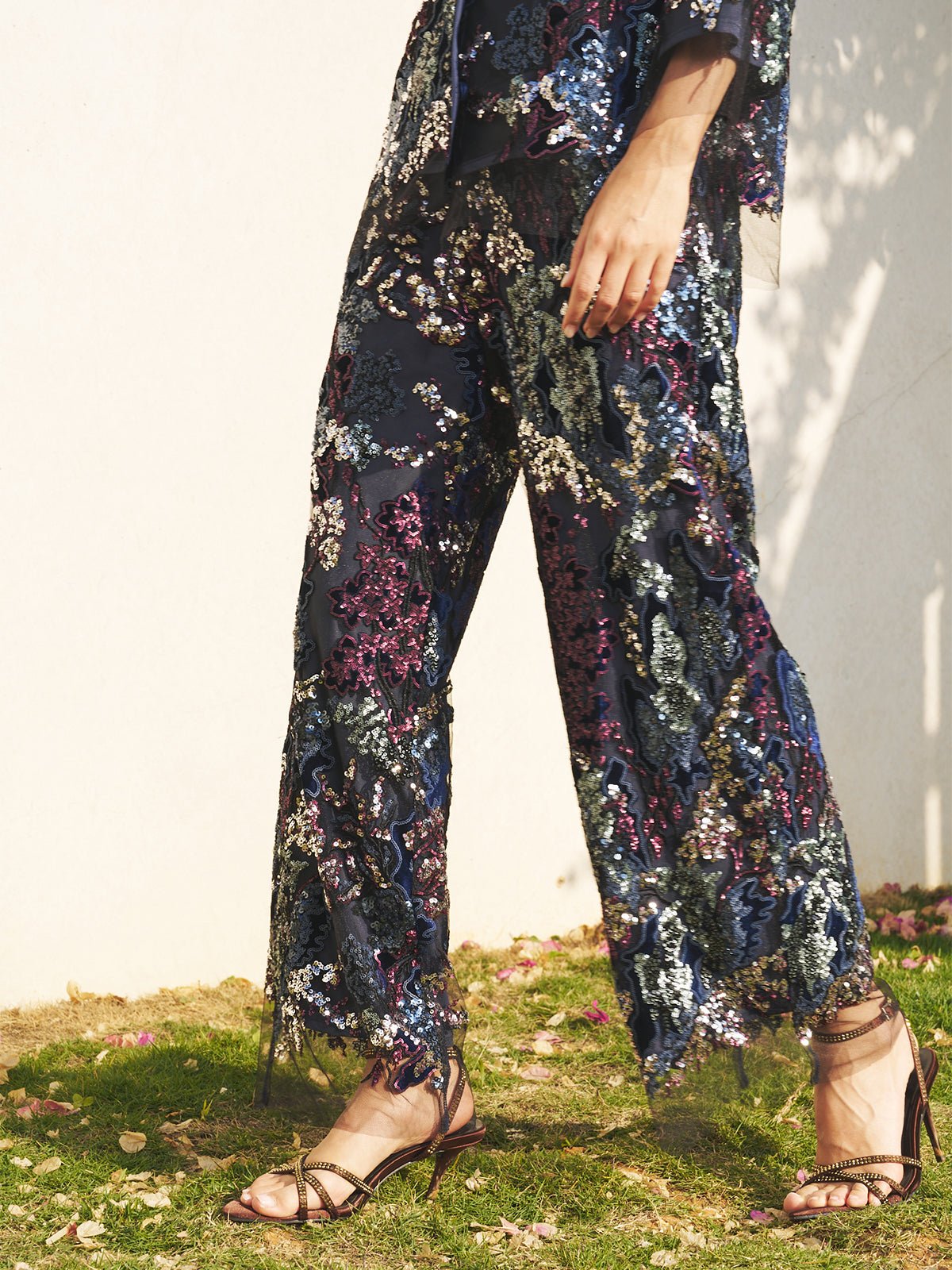 Wearing Social Queen’s Sequin Embroidered Mesh Wide-Leg Pants and strappy sandals, a wedding guest stands on grass by a white wall as light and shadow create an elegant backdrop.