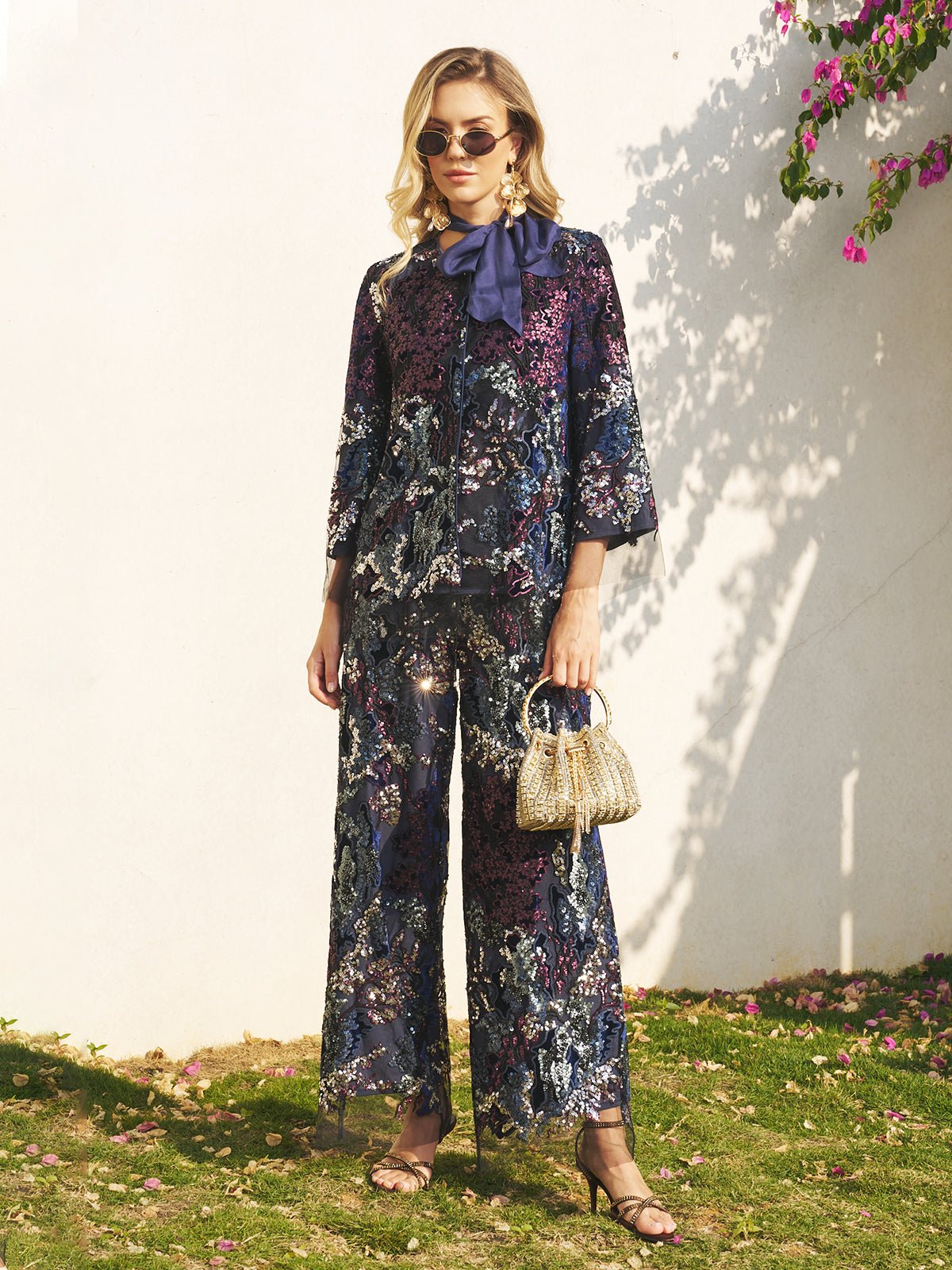 A woman stands on grass in Social Queen’s Sequin Embroidered Mesh Wide-Leg Pants, styled with sunglasses, a large navy bow, heeled sandals, and a beaded handbag—an elegant look for a wedding guest. Pink flowers hang in the top right.