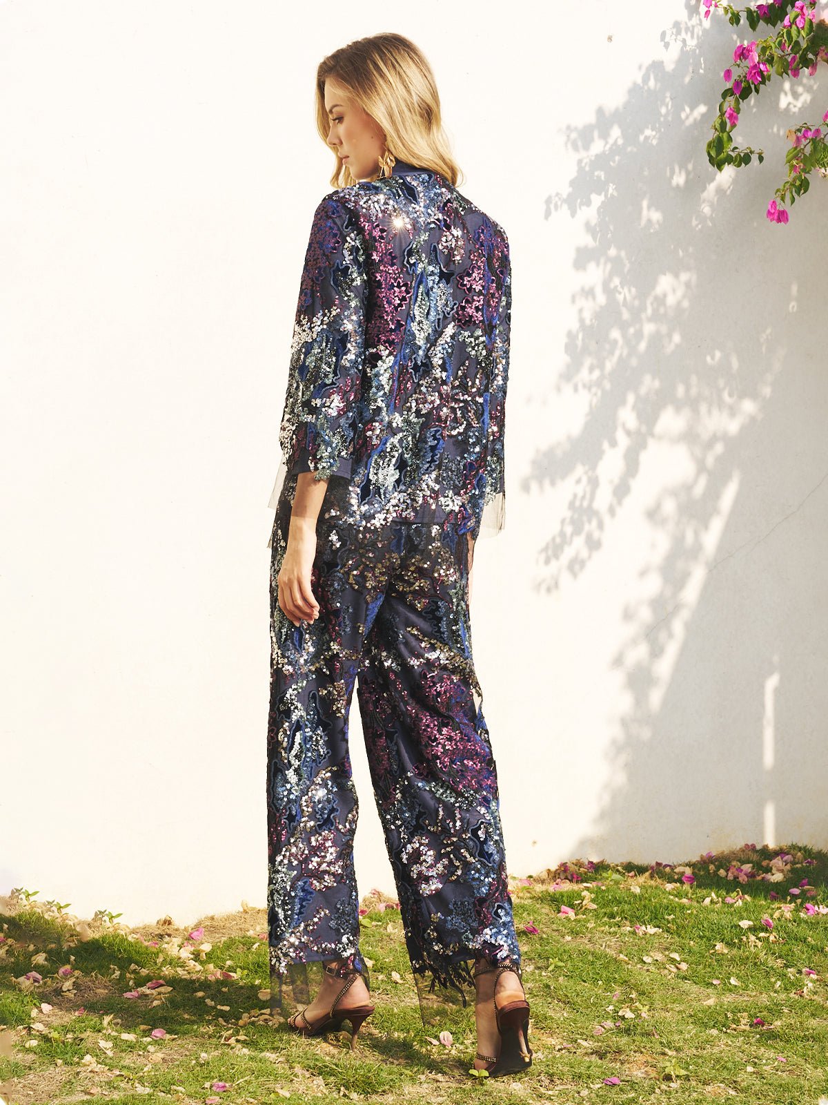 A woman stands on grass near a white wall, wearing Social Queen's Sequin Embroidered Mesh Wide-Leg Pants with heeled sandals—an elegant look perfect for a wedding guest.