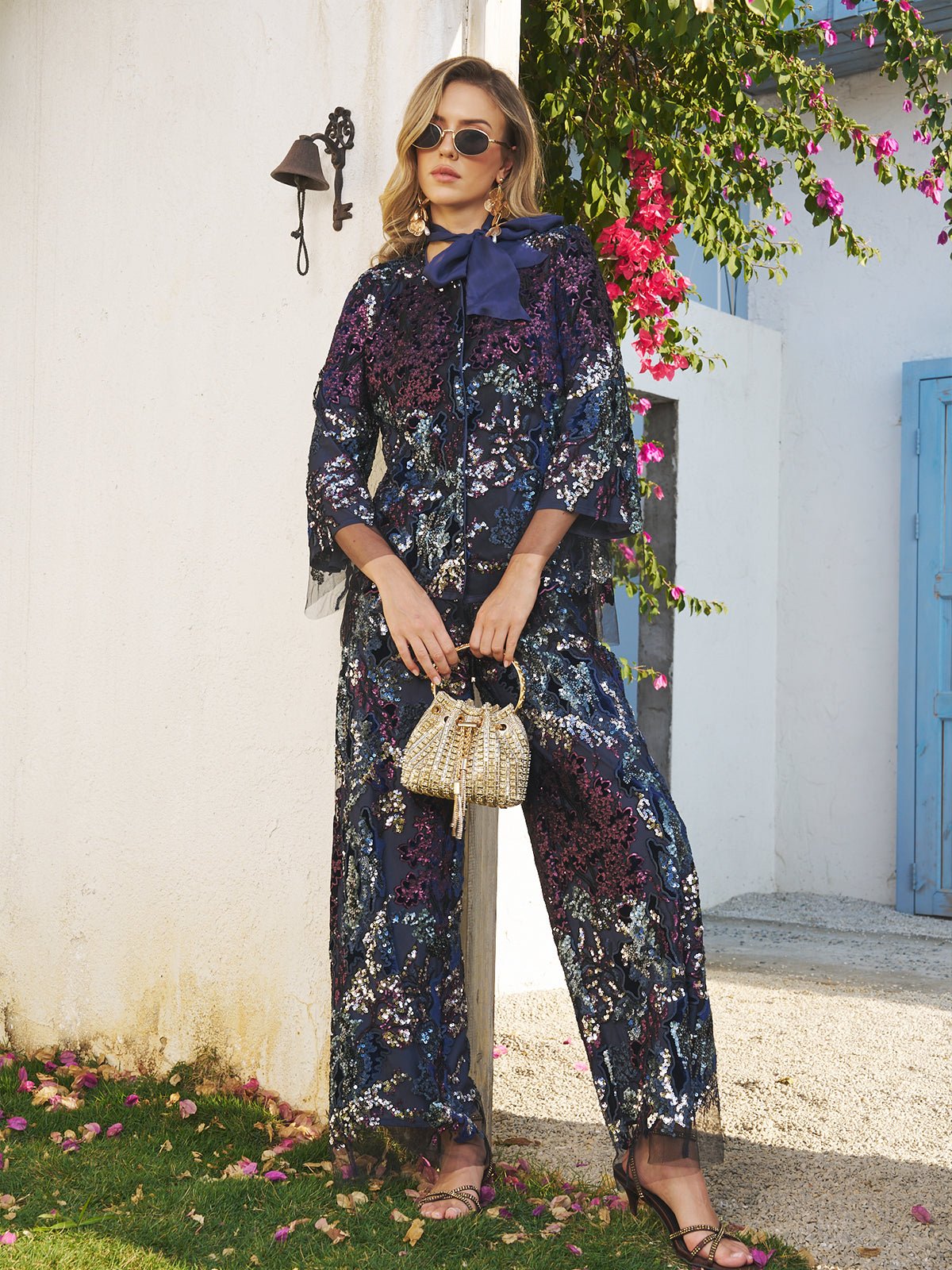 A woman in sunglasses wears the Social Queen Sequin Embroidered Velvet Mesh Jacket, holding a woven handbag while standing outdoors by a white wall with pink flowers and a blue door in the background.