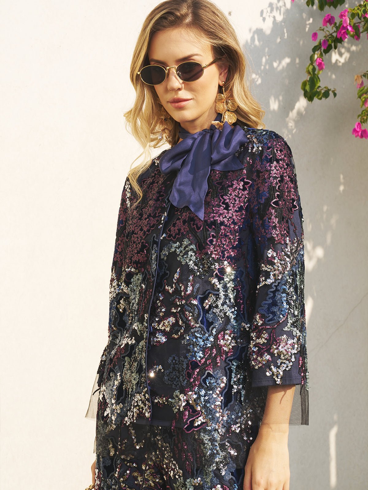 A woman in sunglasses and gold earrings wears the Social Queen Sequin Embroidered Velvet Mesh Jacket with a navy bow, posing against a light wall with pink flowers—perfect for party celebrations.