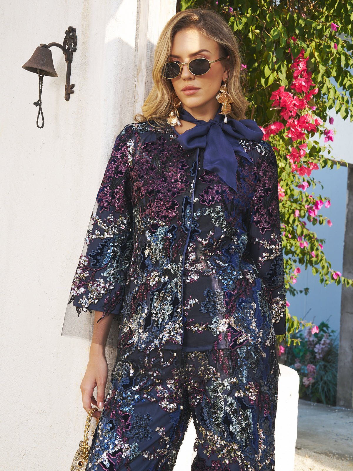A woman wears sunglasses and the Social Queen Sequin Embroidered Velvet Mesh Jacket, featuring navy floral details, with a matching bow, standing outdoors by a white wall and pink flowers.