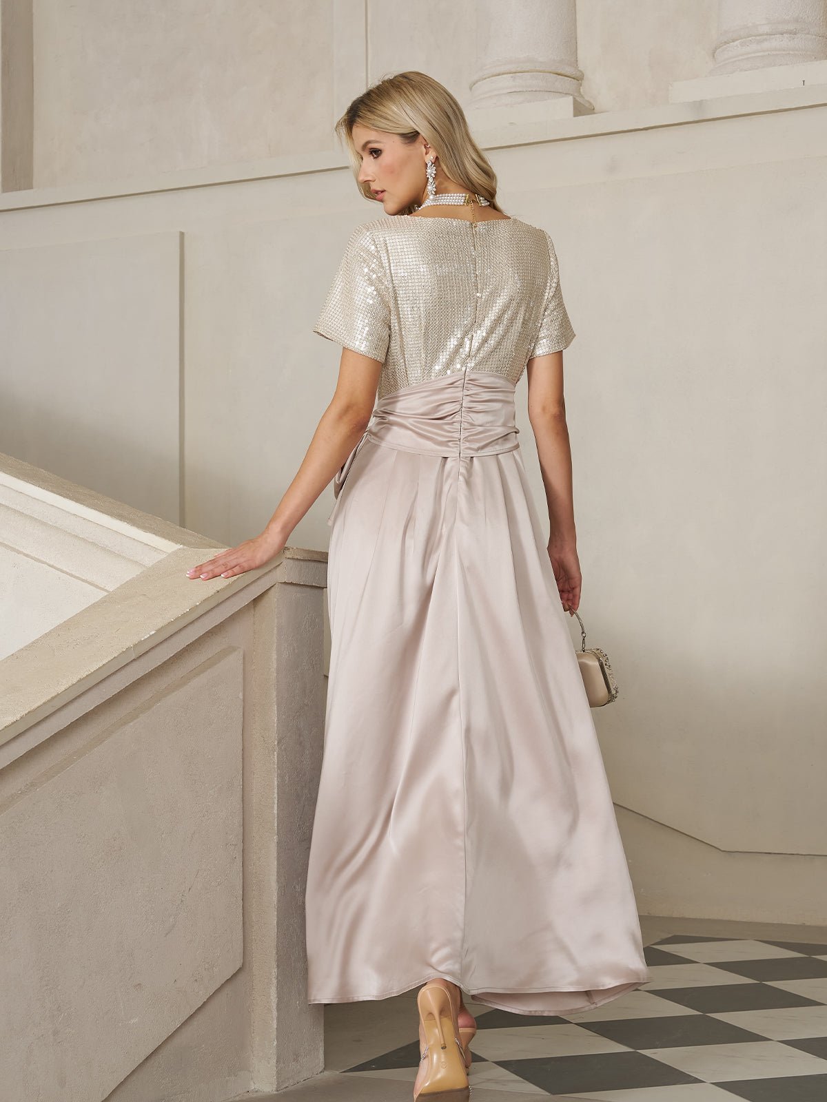 A woman in the Social Queen Sequin & Satin Bow-Waist Dress, featuring a shimmering silver sequin bodice and light pink satin skirt, stands on a staircase facing away, holding a small purse.