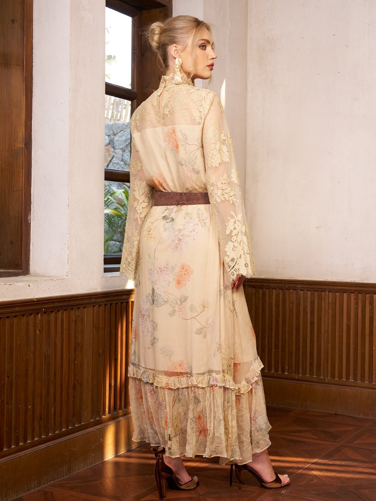 A woman stands indoors near windows, facing away, wearing the Social Queen Silk Floral & Bird Print Lace Dress with long sleeves and a brown belt, paired with brown heels.