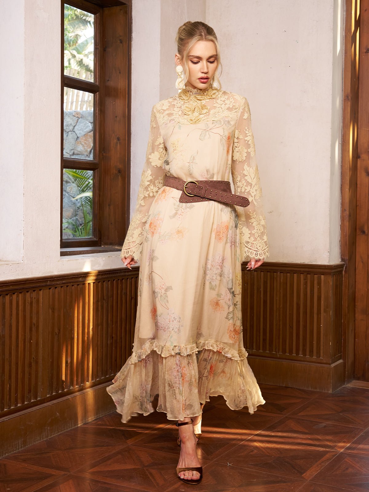 A woman stands indoors wearing the Social Queen Silk Floral & Bird Print Lace Dress, a long light beige piece featuring delicate lace details, paired with a wide brown belt and heeled sandals.