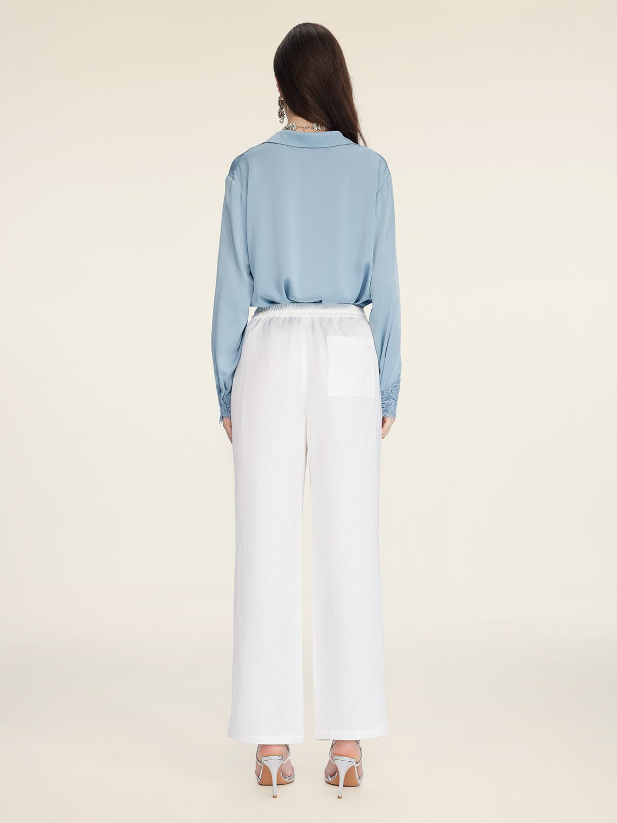 A woman faces away, wearing the Social Queen Silk Satin Shirt with Lace Trim in light blue, paired with white wide-leg pants and white high-heeled sandals against a neutral background.