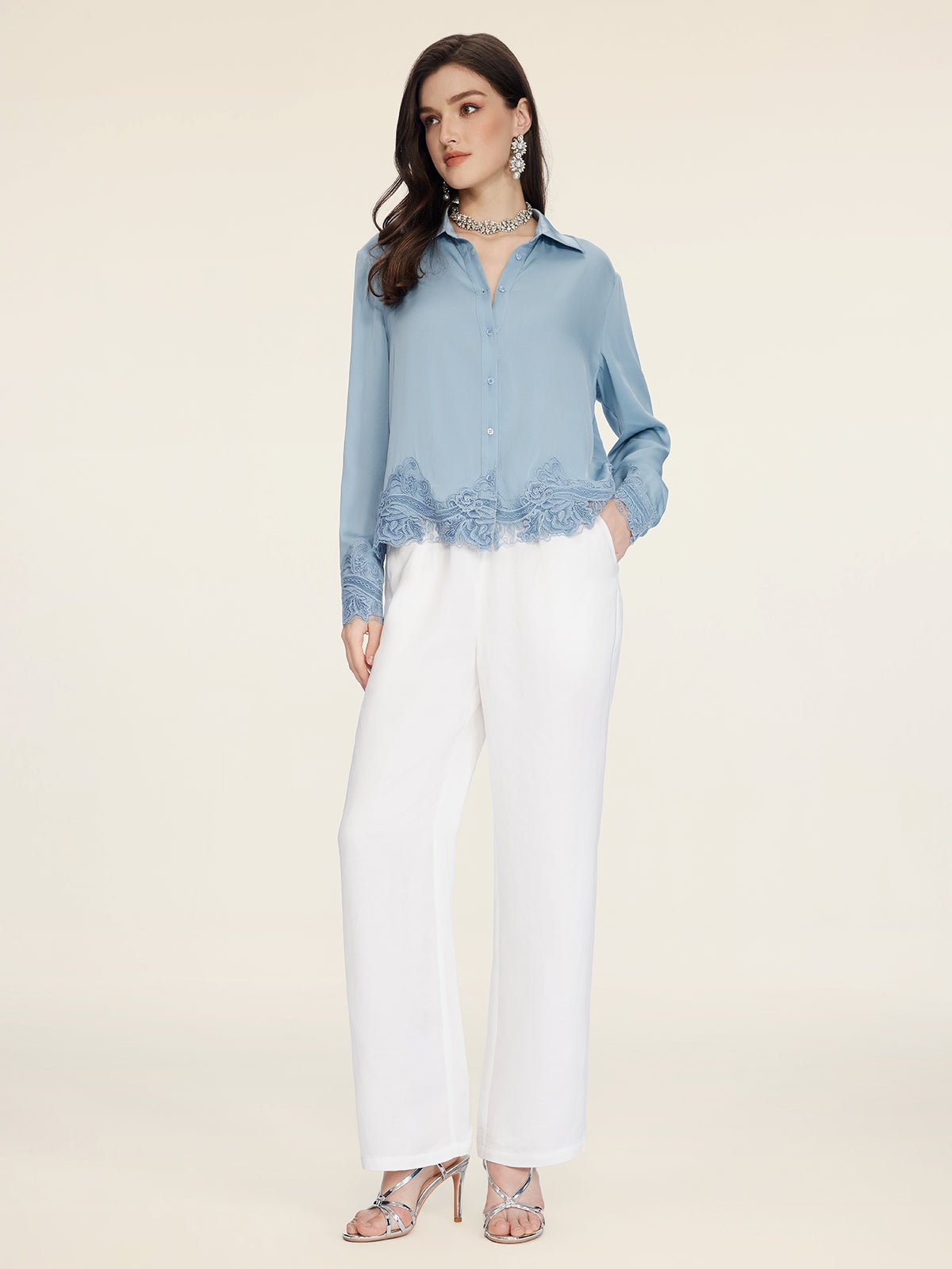 A woman stands against a neutral background wearing the Social Queen Silk Satin Shirt with Lace Trim in light blue, paired with white wide-leg pants, silver heels, and jewelry.