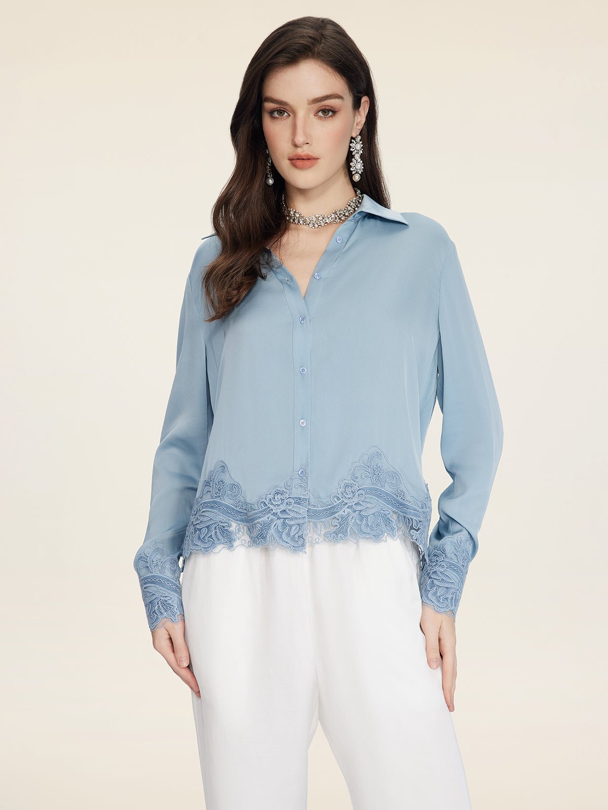 A woman models the Social Queen Silk Satin Shirt with Lace Trim in light blue, featuring long sleeves and hand-stitched lace, paired with white pants against a simple light background.