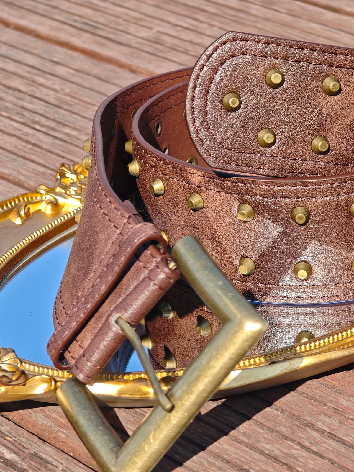 The Social Queen Studded Leather Belt is coiled atop a small oval mirror with a gold frame, displayed on a wooden surface.
