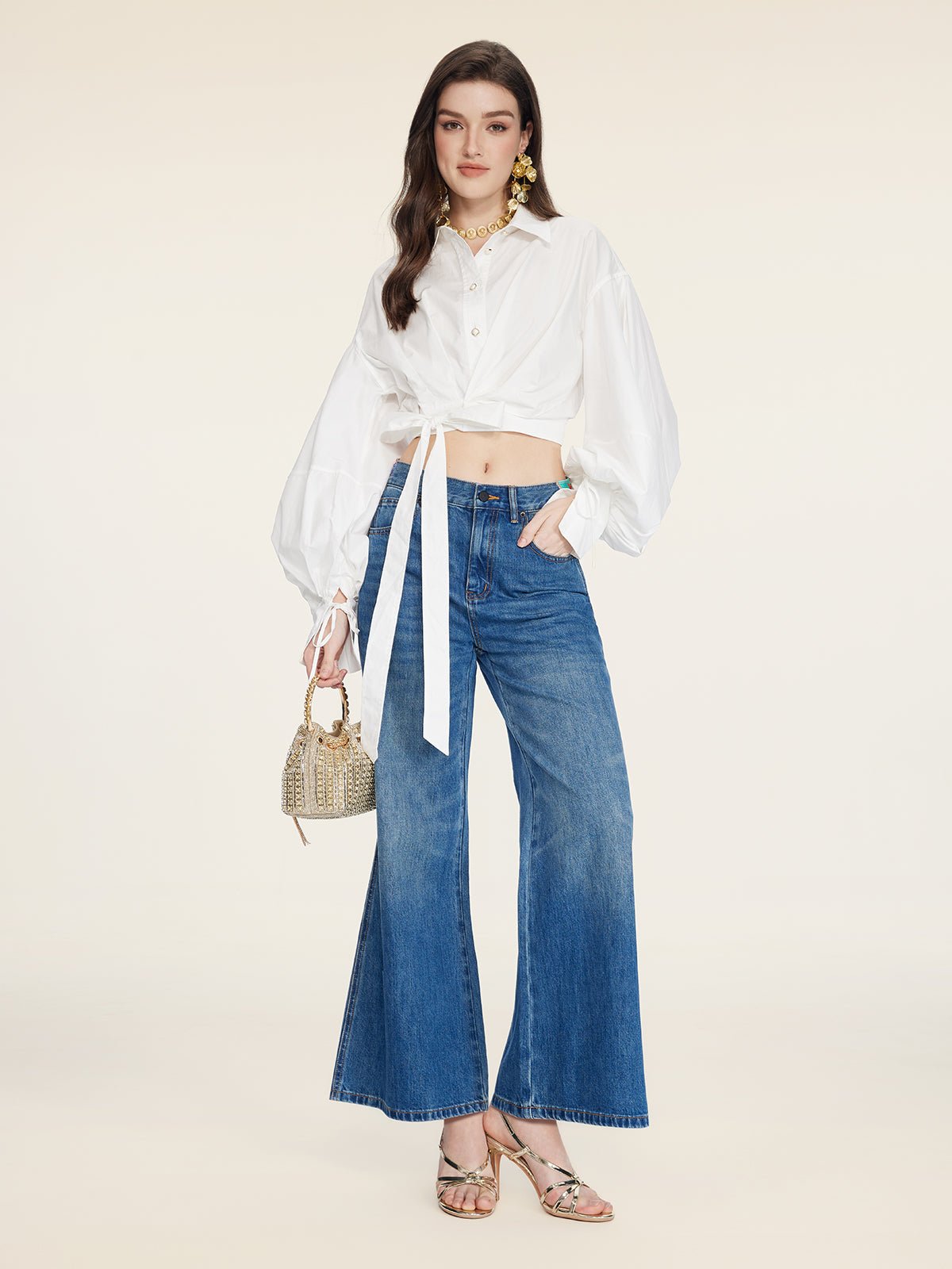 A woman wearing the Social Queen Tie-Neck Silk Lantern Sleeve Blouse, blue wide-leg jeans, heeled sandals, and holding a small woven handbag.
