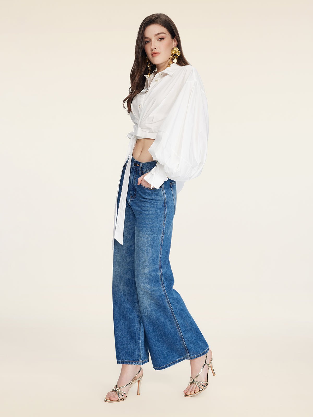 A woman stands sideways against a plain light background, wearing the Social Queen Tie-Neck Silk Lantern Sleeve Blouse, wide-leg blue jeans, and heeled sandals.