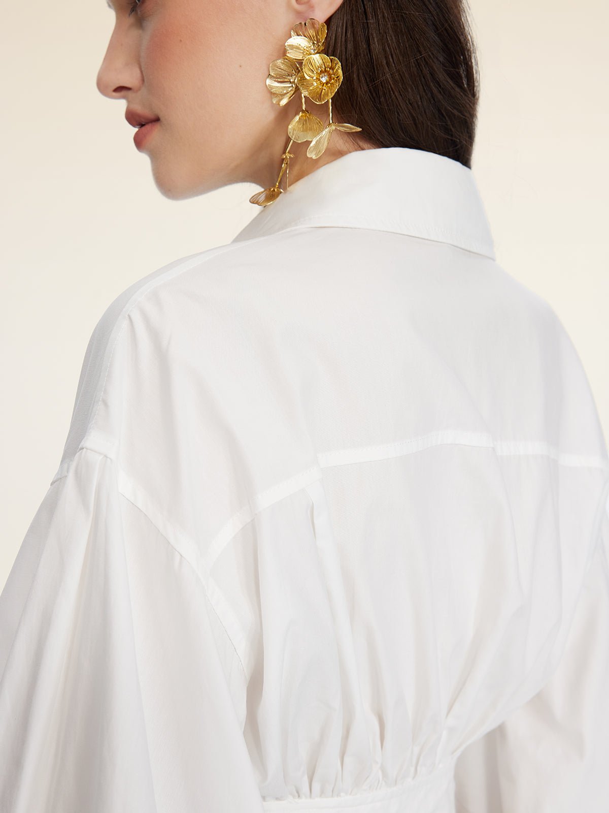 Woman in a white Social Queen Tie-Neck Silk Lantern Sleeve Blouse shown from the back and side, wearing large gold floral earrings with straight brown hair.