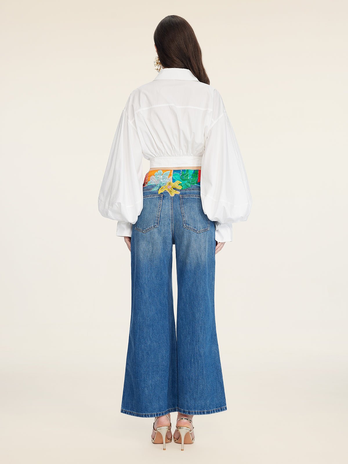 A woman stands facing away, wearing the Social Queen Tie-Neck Silk Lantern Sleeve Blouse in white, paired with wide-leg blue jeans featuring a colorful waistband and heeled sandals.