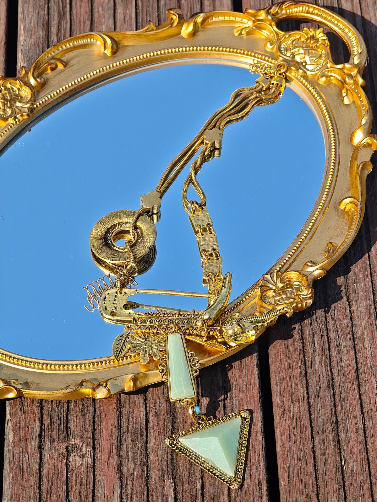 A gold decorative mirror on wooden planks reflects the sky, with Social Queen’s Triangle Pendant Necklace, featuring geometric triangle pendants in artificial jade, resting elegantly on top.