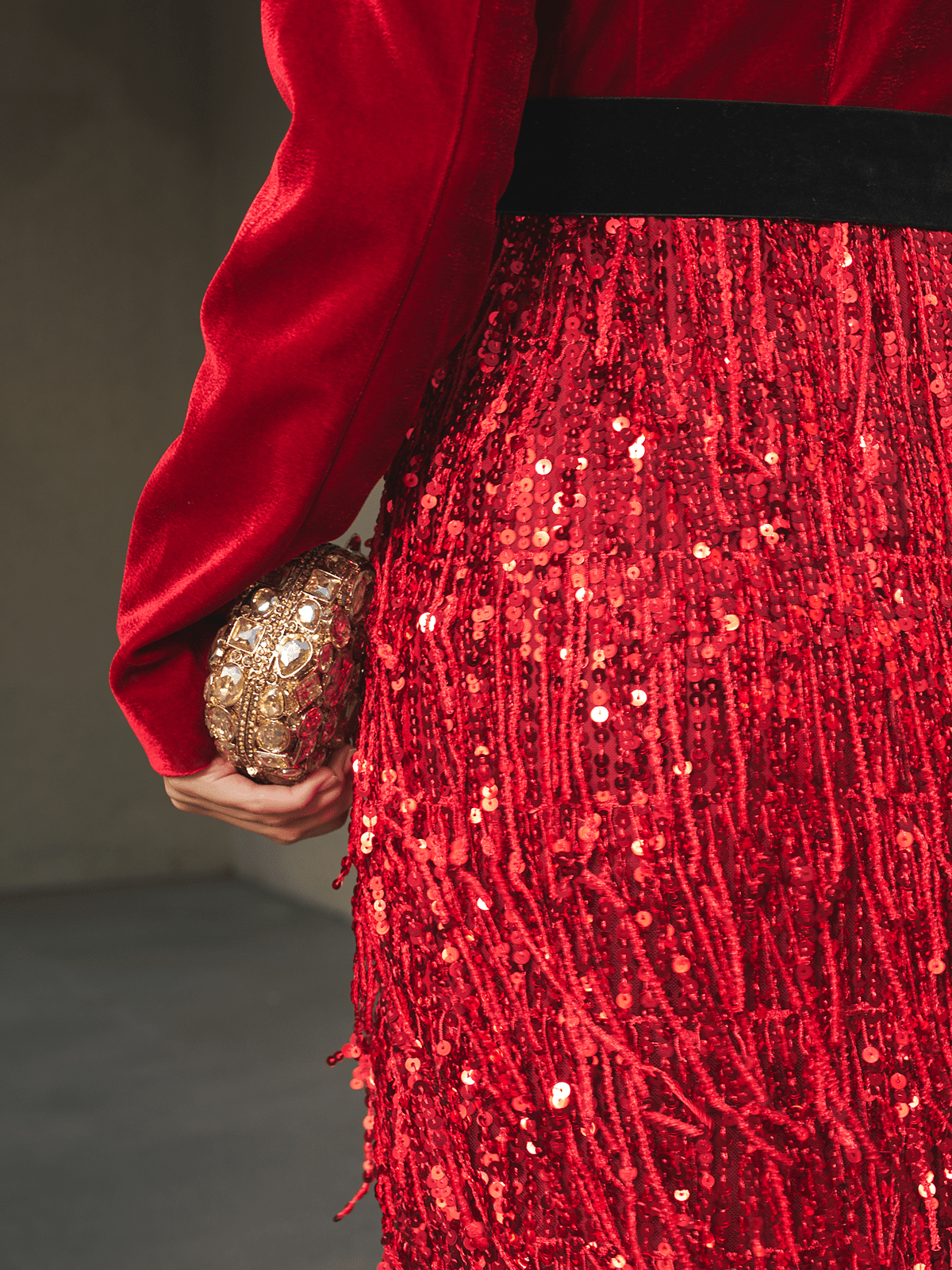 A model in the Social Queen Velvet Fringe Sequin Dress holds a small gold textured clutch in their left hand, set against a softly blurred neutral background.