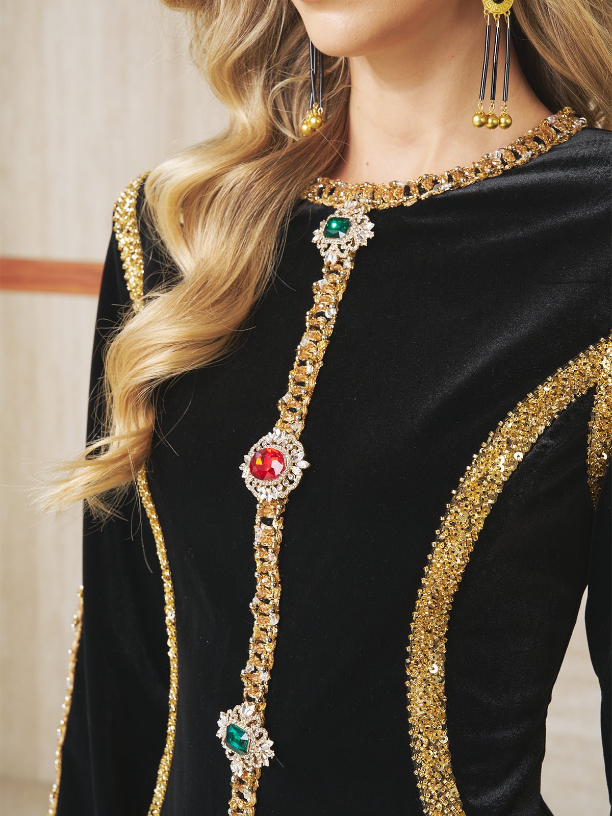 A woman wears the Velvet Jewel-Trim Dress by Social Queen, featuring black velvet with gold sequin trim, jeweled accents, metallic embroidery, and pairs it with long dangling gold earrings.
