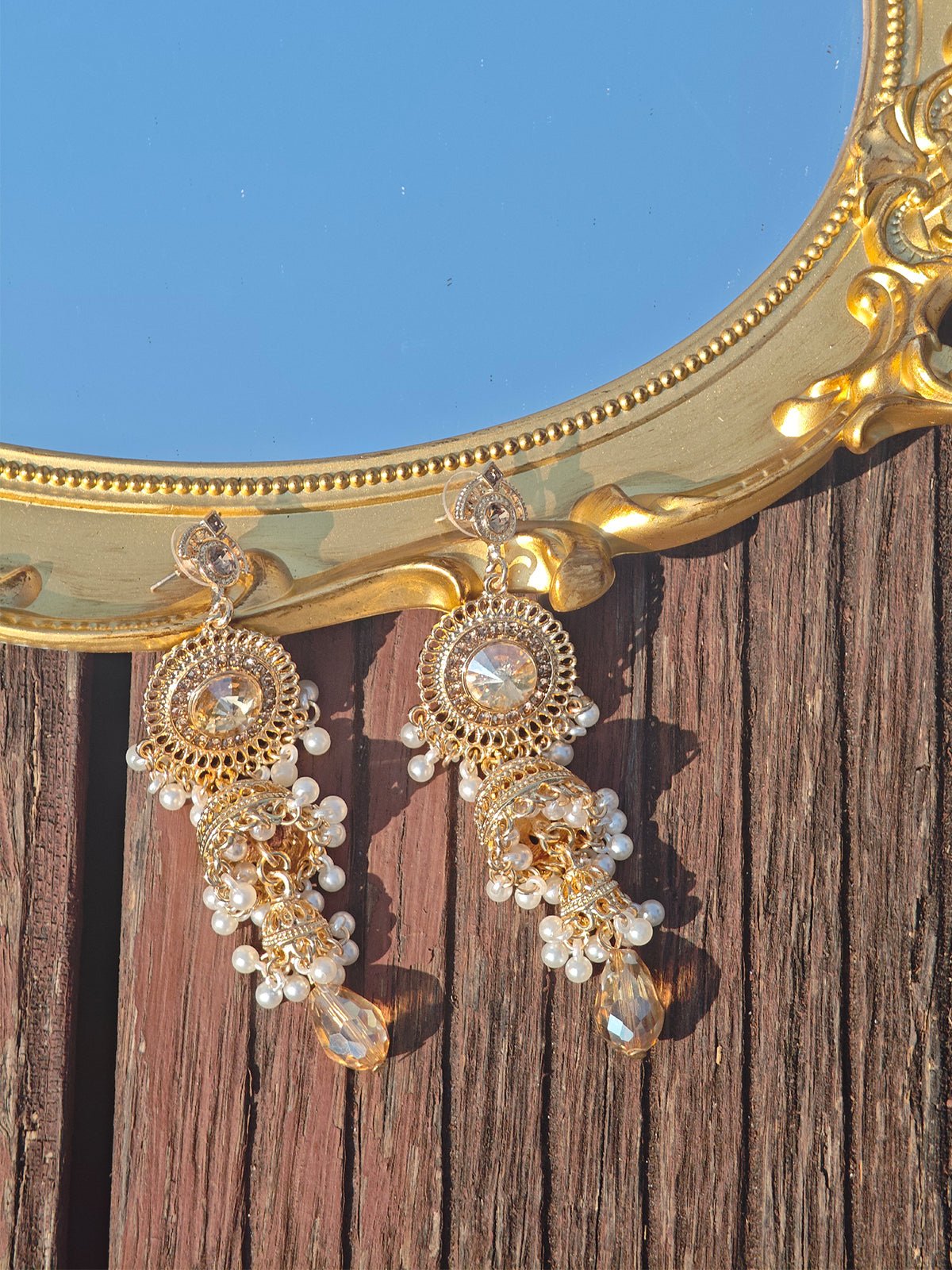 The Vintage Beaded Tassel Earrings by Social Queen feature vintage-inspired pearl tassels with ornate gold and clear gemstones, stylishly displayed on wood beside a decorative gold-framed mirror.