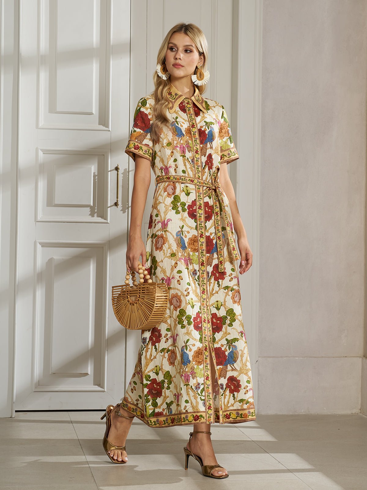 A woman in the Vintage Floral & Bird Print Shirt Dress by Social Queen, paired with a bamboo handbag and gold heeled sandals, stands indoors near white paneled doors—effortlessly chic resort wear.