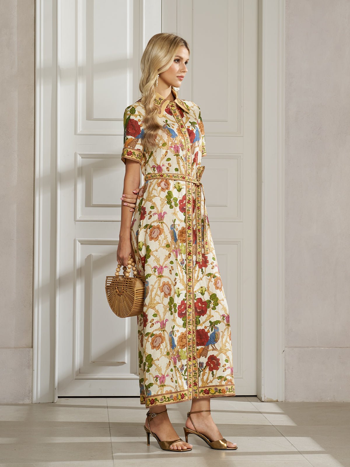 A woman stands indoors wearing the Social Queen Vintage Floral & Bird Print Shirt Dress, accessorized with a wooden handbag and gold heeled sandals—an ideal pick for resort wear. White paneled door and neutral walls set a chic backdrop.