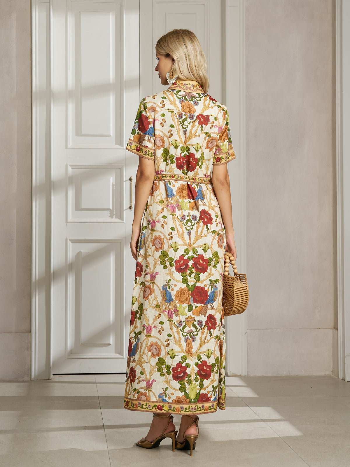 A woman in the Social Queen Vintage Floral & Bird Print Shirt Dress stands indoors by a white door, holding a woven handbag and wearing heeled sandals—an elegant resort-ready look.
