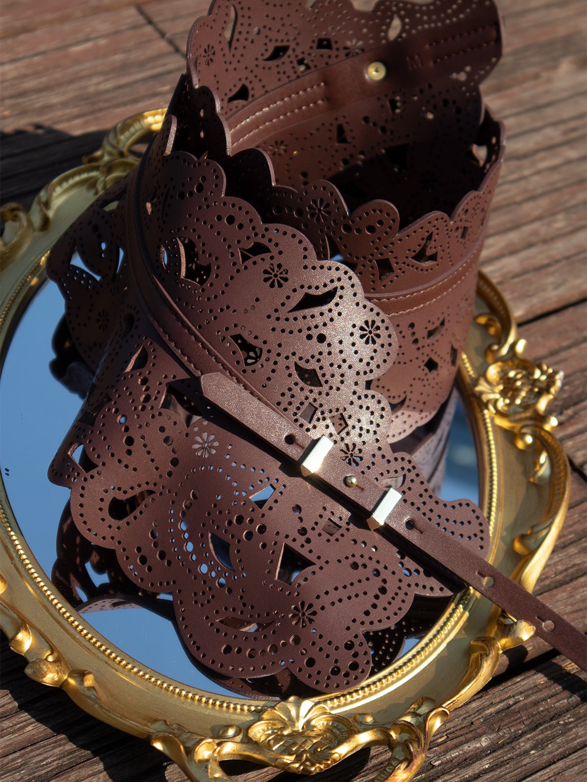 The Vintage Floral Cutout Waist Belt by Social Queen, featuring intricate laser-cut details and a scalloped edge in top grain leather, is showcased on an ornate gold-framed mirror atop wooden planks.