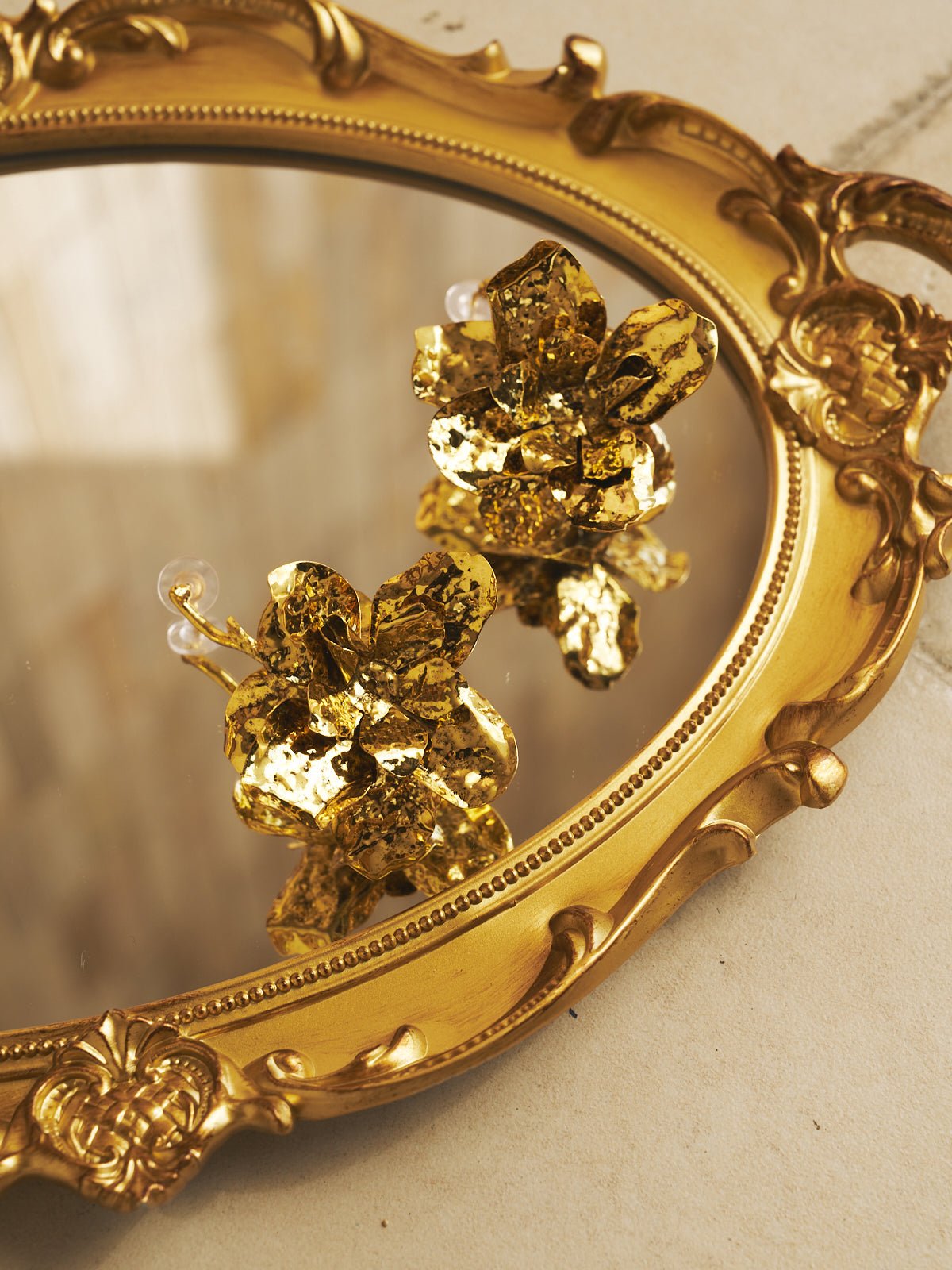 The Vintage Floral Earrings by Social Queen are displayed on an ornate gold-toned oval mirror, their floral design reflected elegantly against a light background.