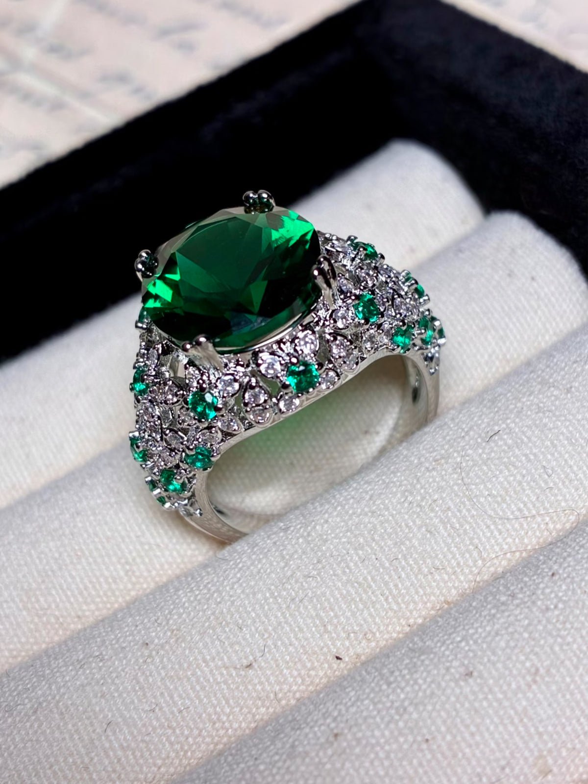 The Social Queen Vintage Green Crystal Ring features a large oval green gemstone accented with clear and green stones, white gold plated, and comes elegantly displayed in a jewelry box.