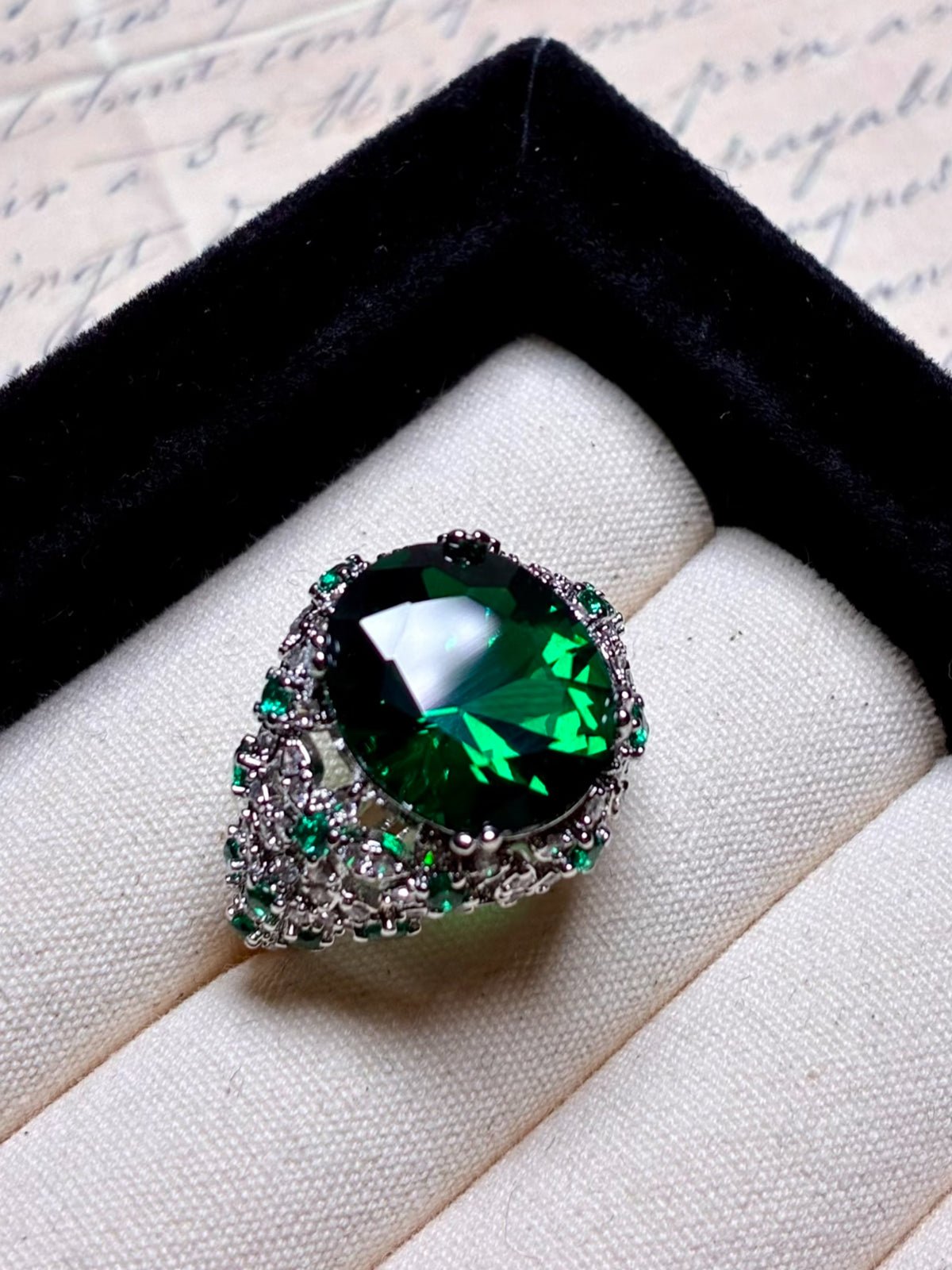 The Social Queen Vintage Green Crystal Ring features an oval-cut green gemstone with smaller green and clear stones, a white gold plated adjustable open band, and comes elegantly displayed in a jewelry box.