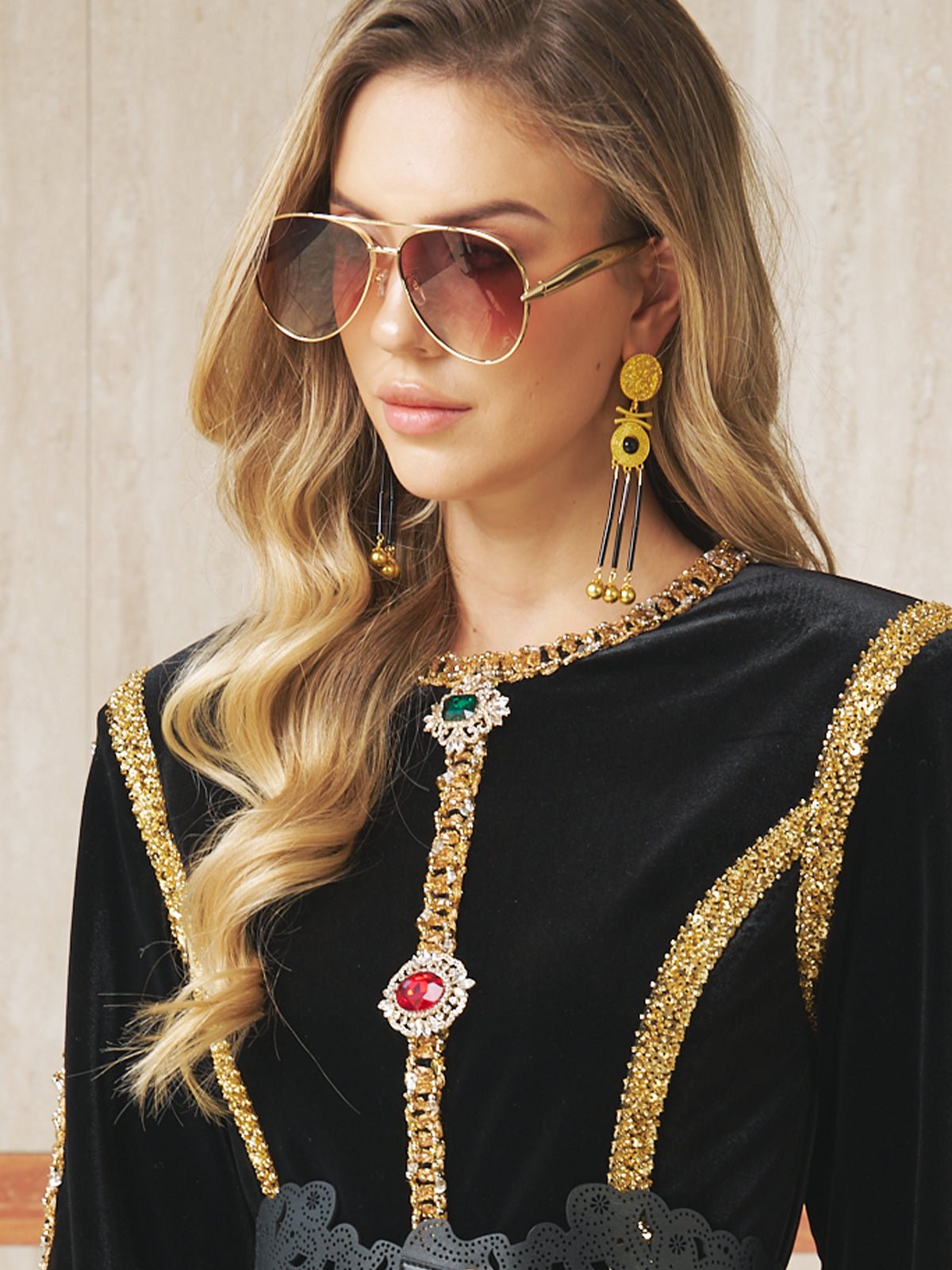A woman in Social Queen’s Vintage Sunglasses with Gold Frame, a black and gold embellished dress, and statement earrings stands against a neutral background.