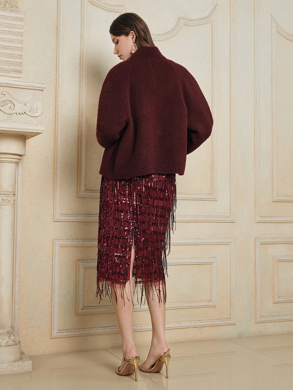 A woman stands with her back to the camera, wearing Social Queen's Wool-Cashmere Cropped Wrap Jacket in burgundy over a red sequin fringe skirt and gold heels, set against ornate cream-colored walls.