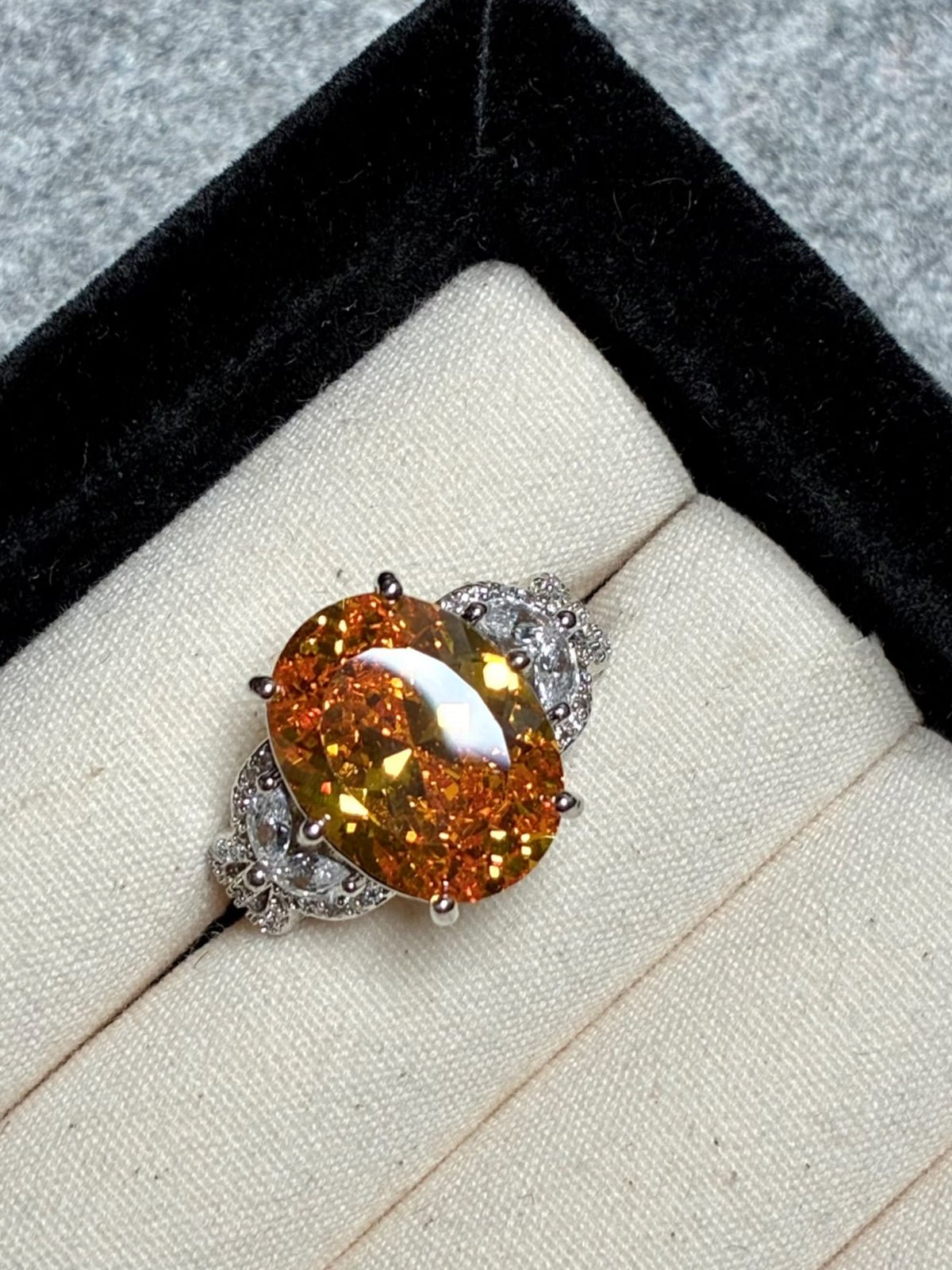 The Social Queen Yellow Crystal Ring features a gold-orange oval crystal with diamond accents on an adjustable white gold band, presented in a black and cream jewelry box—a striking statement piece.