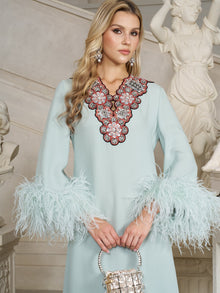 A woman stands indoors among classical statues, holding a silver bag and wearing the Social Queen 100% Mulberry Silk Feather Limited Dress, featuring ostrich feather sleeves and an ornate beaded neckline in light blue.