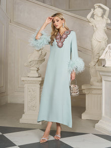 A woman in the Social Queen 100% Mulberry Silk Feather Limited Dress, with ostrich feather sleeves and an embroidered neckline, stands indoors by classical statues holding a small handbag.