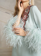 A person models the Social Queen 100% Mulberry Silk Feather Limited Dress, a vintage-inspired light blue evening gown featuring ostrich feather sleeves and ornate beaded embellishments, with one hand touching their face.