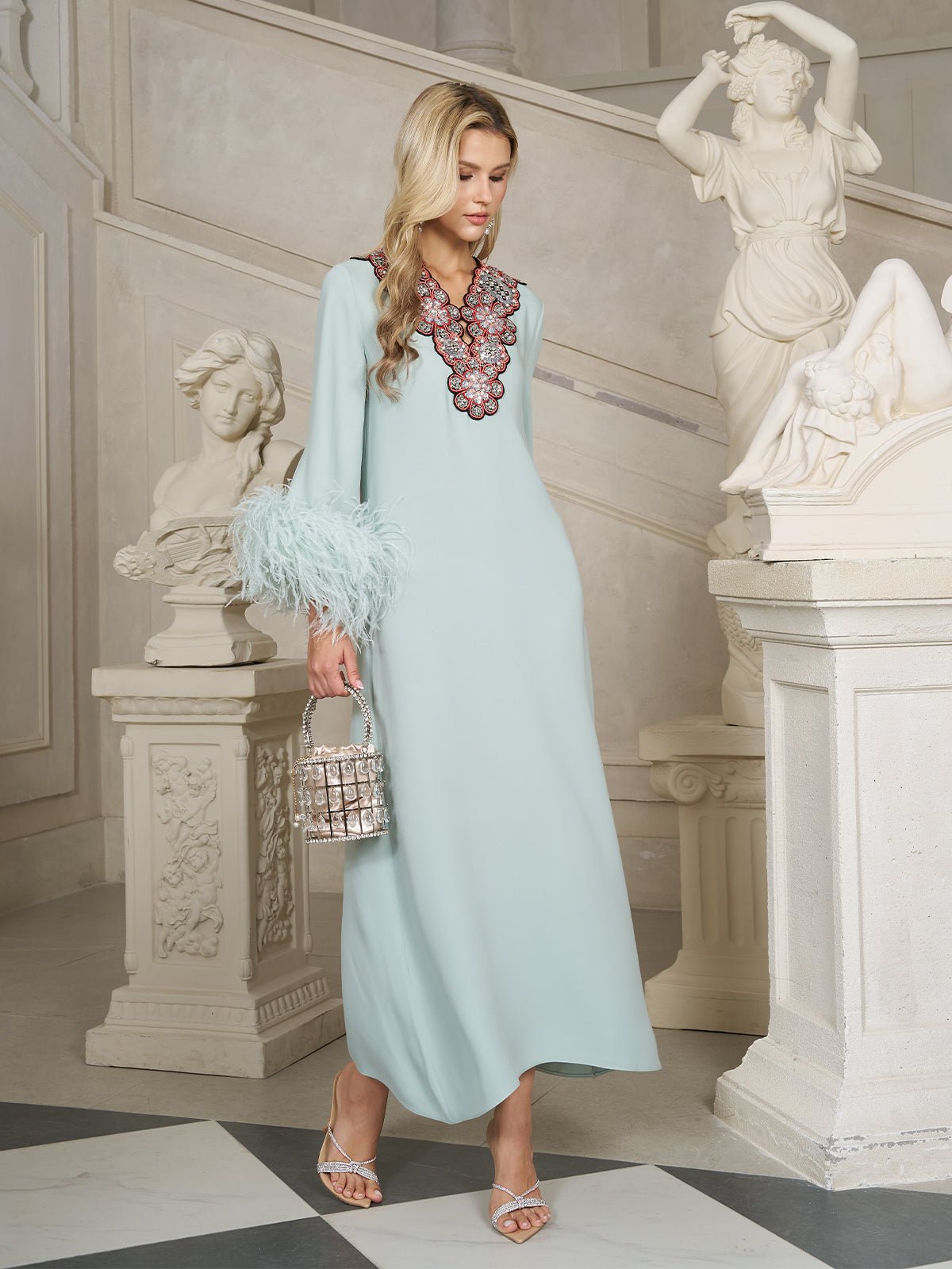 A woman in the Social Queen 100% Mulberry Silk Feather Limited Dress, featuring ostrich feather sleeves and an embellished neckline, walks across marble floors with classical statues, carrying a silver handbag.