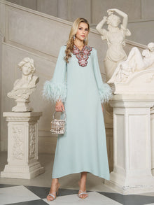 A woman wearing the Social Queen 100% Mulberry Silk Feather Limited Dress with ostrich feather sleeves and a jeweled neckline stands indoors by white classical statues, holding a small handbag.