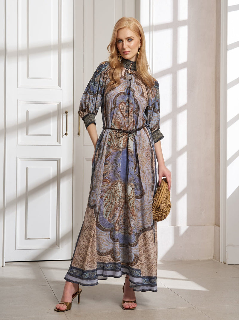 A woman stands indoors in the Social Queen 100% Mulberry Silk Paisley Printed Dress with an A-line silhouette, belted waist, holding a woven clutch and wearing heels near a sunlit white door and window.