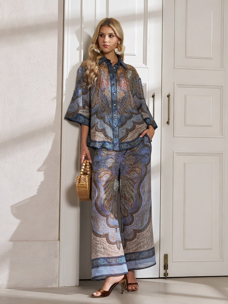 A woman stands indoors wearing Social Queen’s 100% Mulberry Silk Paisley Printed Trousers, paired with a vintage shirt, a woven handbag, and heeled sandals.