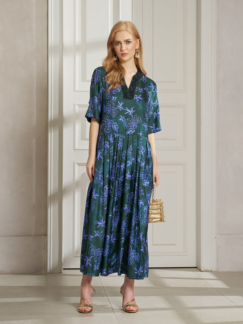 A woman stands indoors wearing the Social Queen 100% Mulberry Silk Printed Beaded Dress, paired with gold sandals, statement earrings, and a small gold purse.