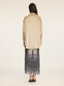 A person stands facing away, wearing the Social Queen 100% Silk Embroidered Lace Satin Blouse in beige, paired with a sequined silver fringe skirt and strappy heels against a plain light background.