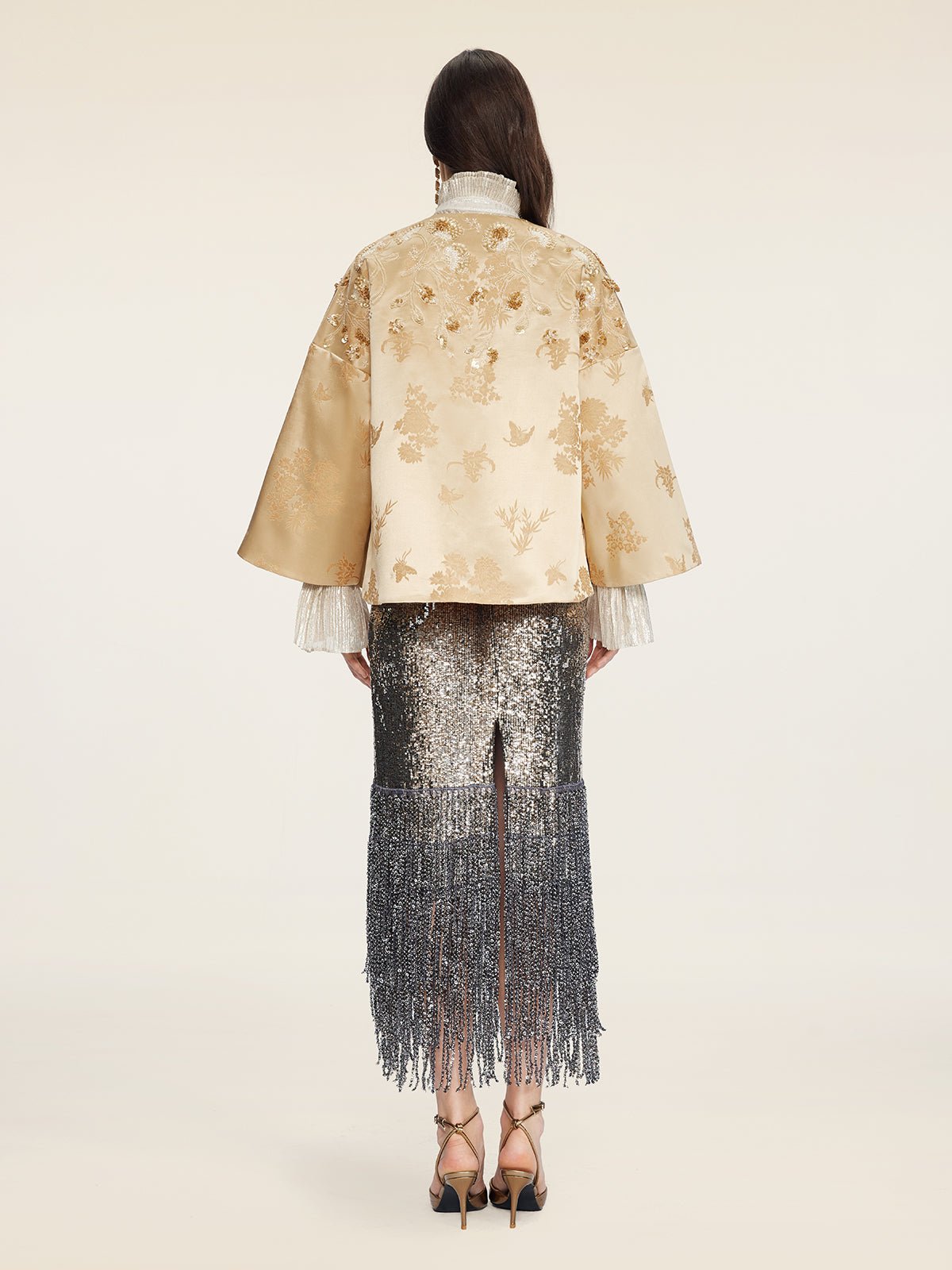 A person stands facing away, wearing the Social Queen 100% Silk Floral Embroidered Satin Jacket with wide sleeves, paired with a silver sequin fringe skirt, heels, and earrings against a plain background.