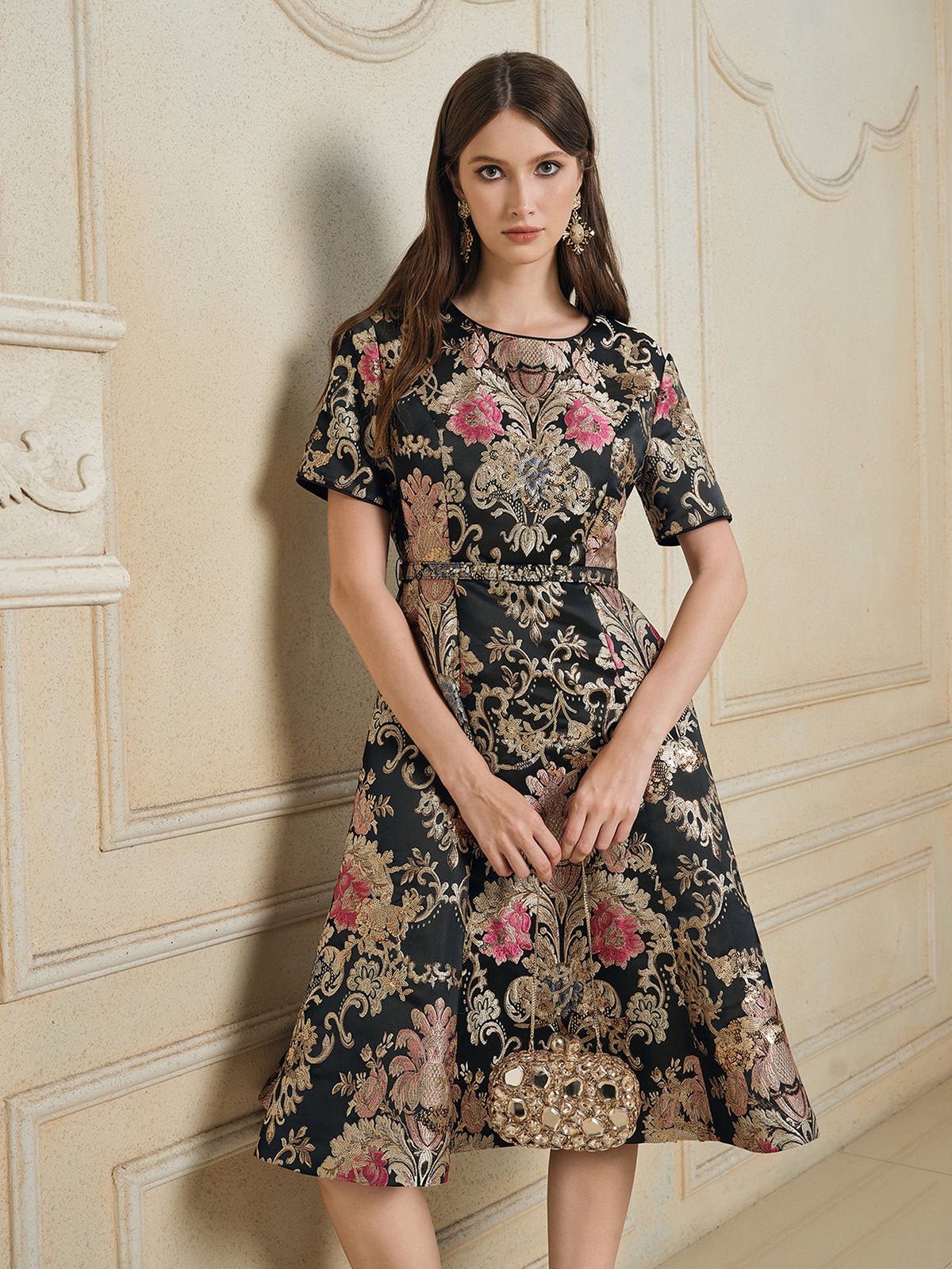 A woman stands indoors in the Social Queen 100% Silk Jacquard Satin Gold Embroidered Floral Dress, holding a decorative handbag against a beige ornate wall.