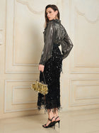 A woman stands indoors, wearing the Social Queen 100% Silk Luminous Metallic Ruffle Blouse, a black sequin fringe skirt, black heels, and holding a gold handbag. She faces sideways against a decorative wall.