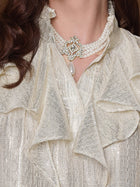 A woman wears the Social Queen 100% Silk Luminous Metallic Ruffle Blouse with puff sleeves and a ruffled neckline, paired with a multi-strand pearl choker necklace featuring a jeweled pendant and a large pearl drop.