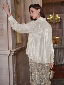 A woman in a Social Queen 100% Silk Luminous Metallic Ruffle Blouse with puff sleeves and a sequin skirt stands by an ornate wall, holding a small clutch, with a chandelier visible in the background.