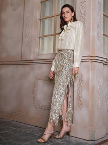 A woman stands indoors against an ornate wall, wearing the Social Queen 100% Silk Luminous Metallic Ruffle Blouse paired with a silver sequin fringed skirt and strappy heels.