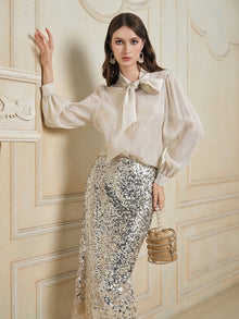 A woman wears the Social Queen 100% Silk Metallic Ribbon-Tie Blouse with a silver sequin skirt and holds a small handbag, standing against an ornate beige wall.