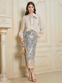 Indoors, a woman wears the Social Queen 100% Silk Metallic Ribbon-Tie Blouse with puff sleeves and a large bow, silver sequin midi skirt, gold sandals, and holds a gold handbag.