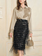 A woman wears the Social Queen 100% Silk Retro Floral Appliqué Satin Blouse, tucked into a black sequined fringe skirt, holding a beaded handbag while standing against a cream-colored wall.