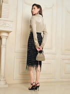 A woman stands indoors facing away, exuding French vintage elegance in a Social Queen 100% Silk Retro Floral Appliqué Satin Blouse, paired with a black fringe skirt, black heels, and a small beige handbag.
